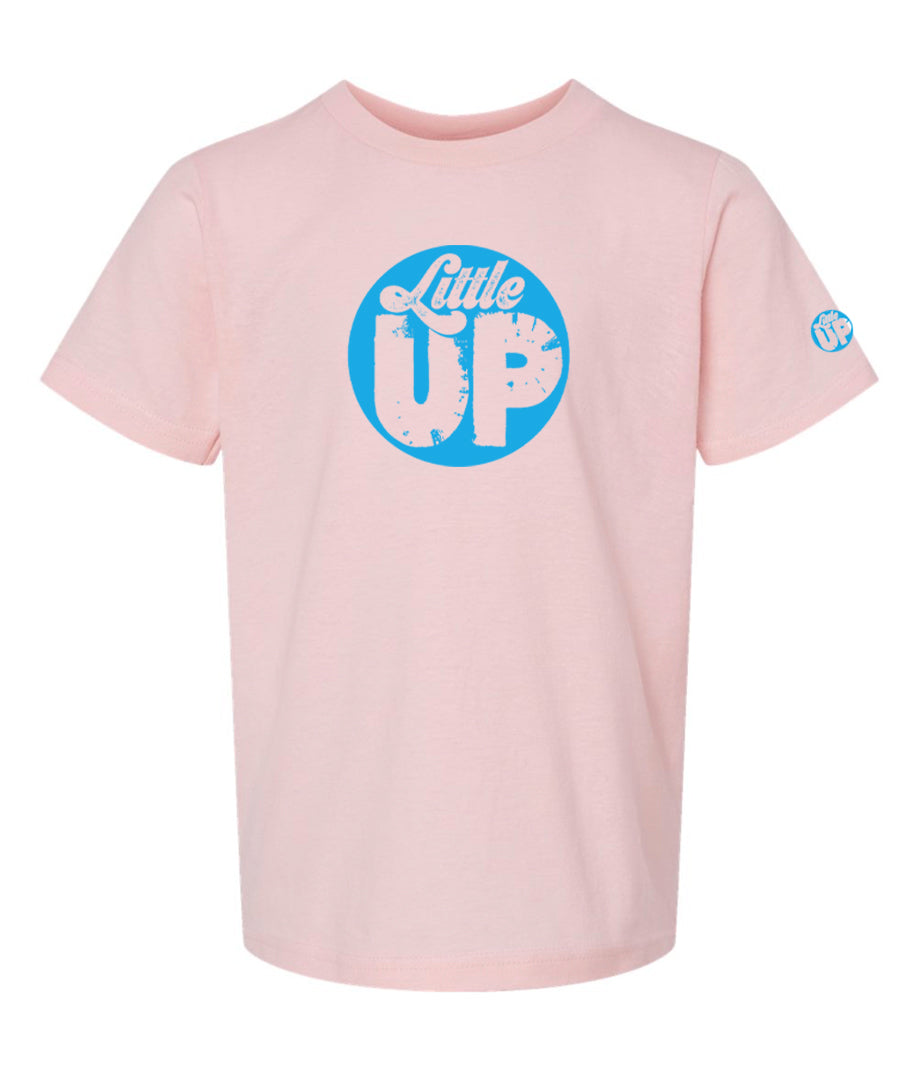 light blue circle logo with "Little" in the top third in textured seventies script and "UP" on the bottom ⅔ in modern sans serif textured font on pink youth t-shirt on white studio background