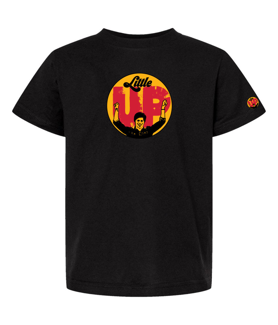 large circle with "Little" at the top in 70s script, "UP" large in red textured sans serif, illustration of young man smiling with arms raised illustration in black at the bottom, on black youth t-shirt