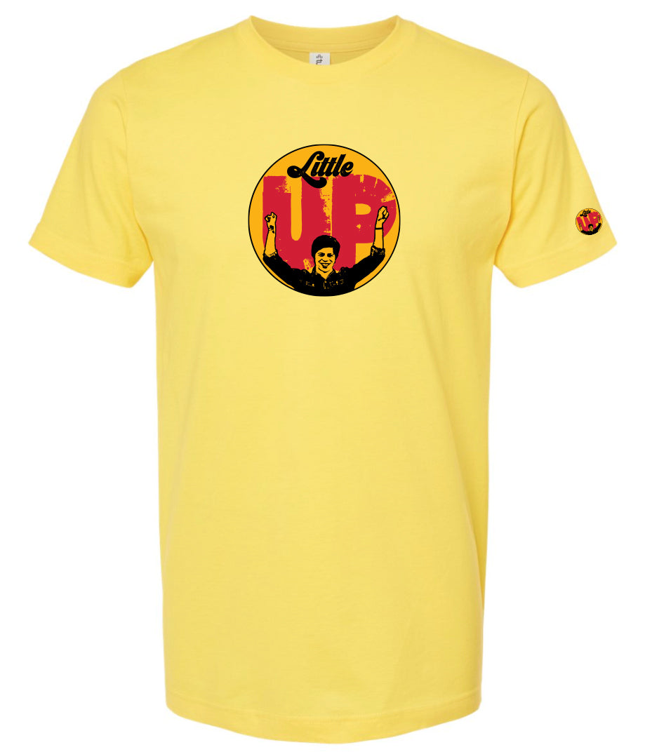 large circle with "Little" at the top in 70s script, "UP" large in red textured sans serif, illustration of young man smiling with arms raised illustration in black at the bottom, on yellow t-shirt